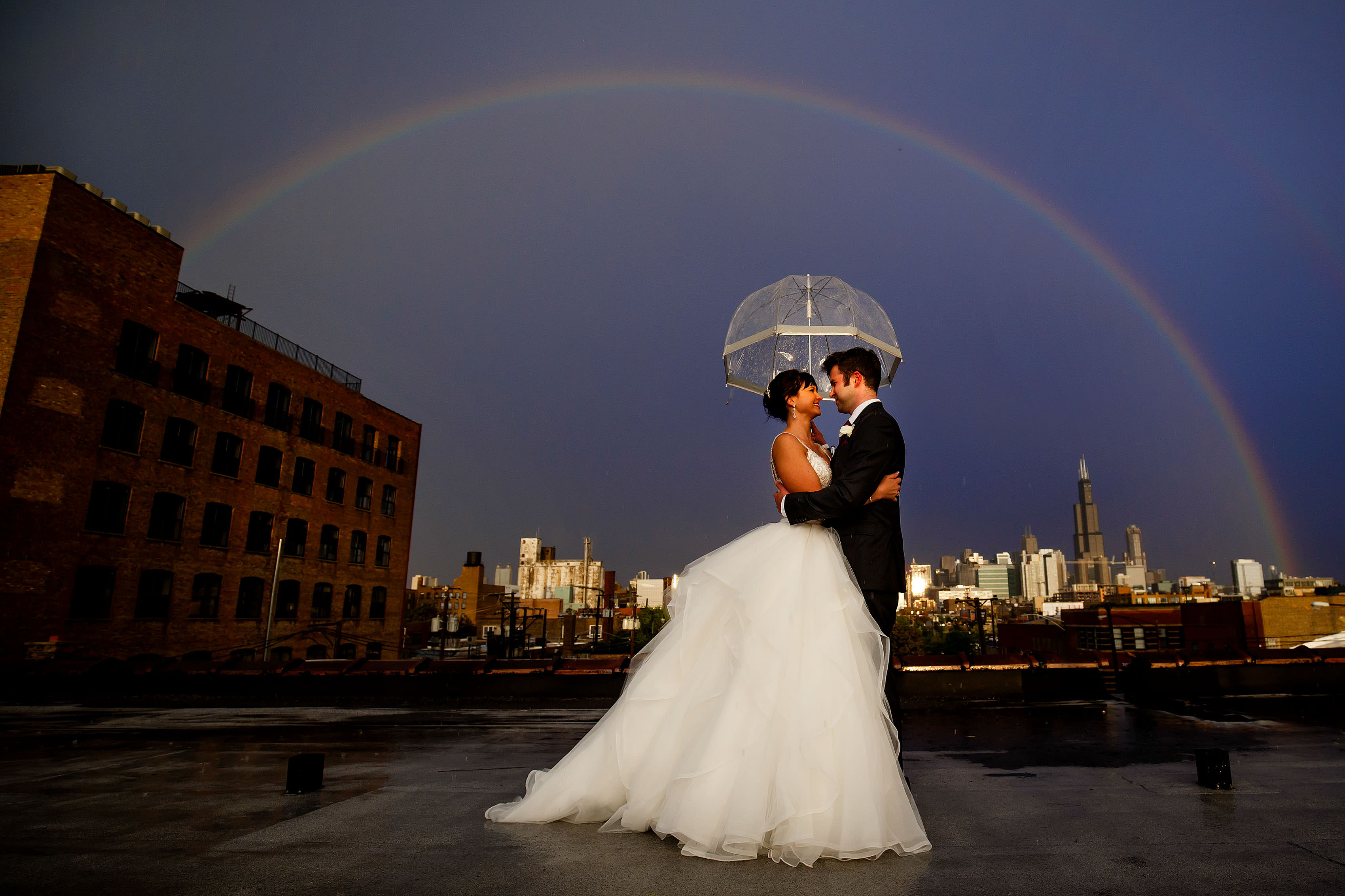 The bride and Groom embrace under an umbrella and surrounded by a rainbow on the rooftop during their Room 1520 wedding in Chicago