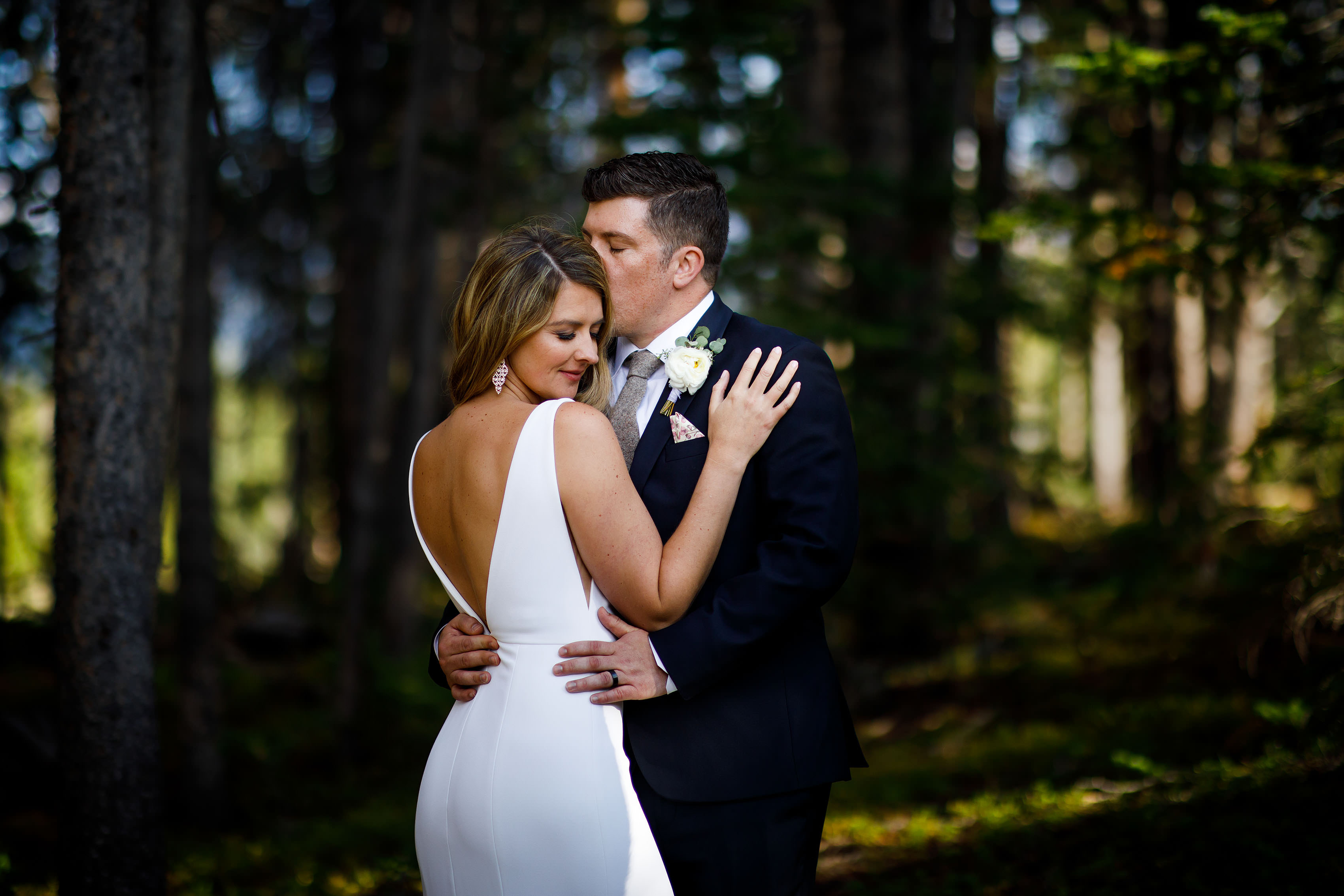 The bride poses while the groom gives her a kiss in a backless sexy gown  during their wedding at TenMile Station - Justin Edmonds Photography