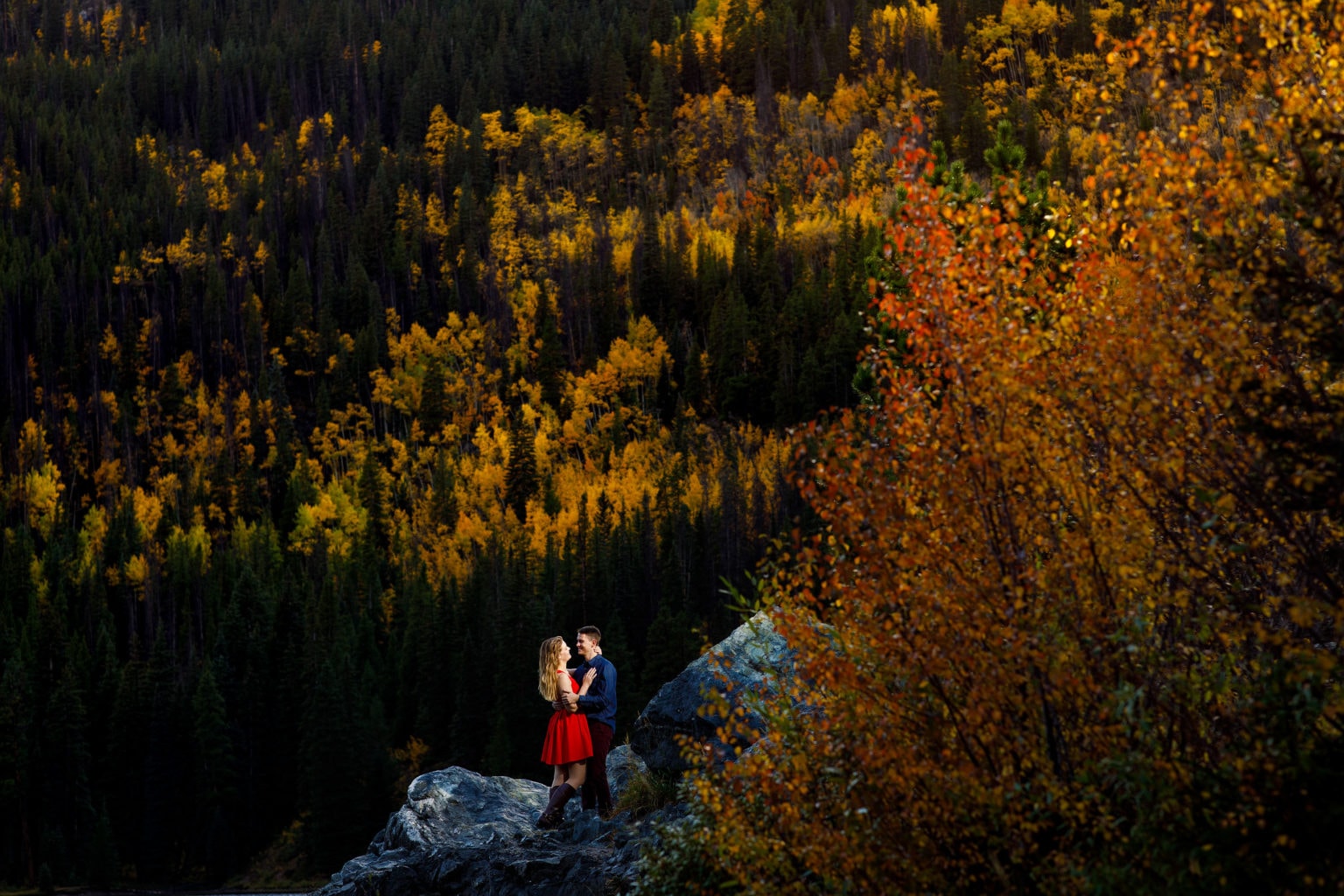 A couple pose on a rock surrounded by golden fall colors at Officers Gulch during their fall mountain engagement photos near Frisco Colorado