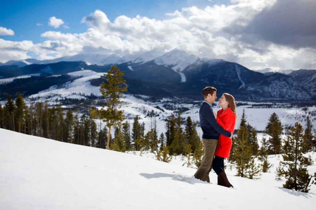 Jessica wears a red coat while embracing with Mike in the snow at Sapphire Point during their winter keystone engagement
