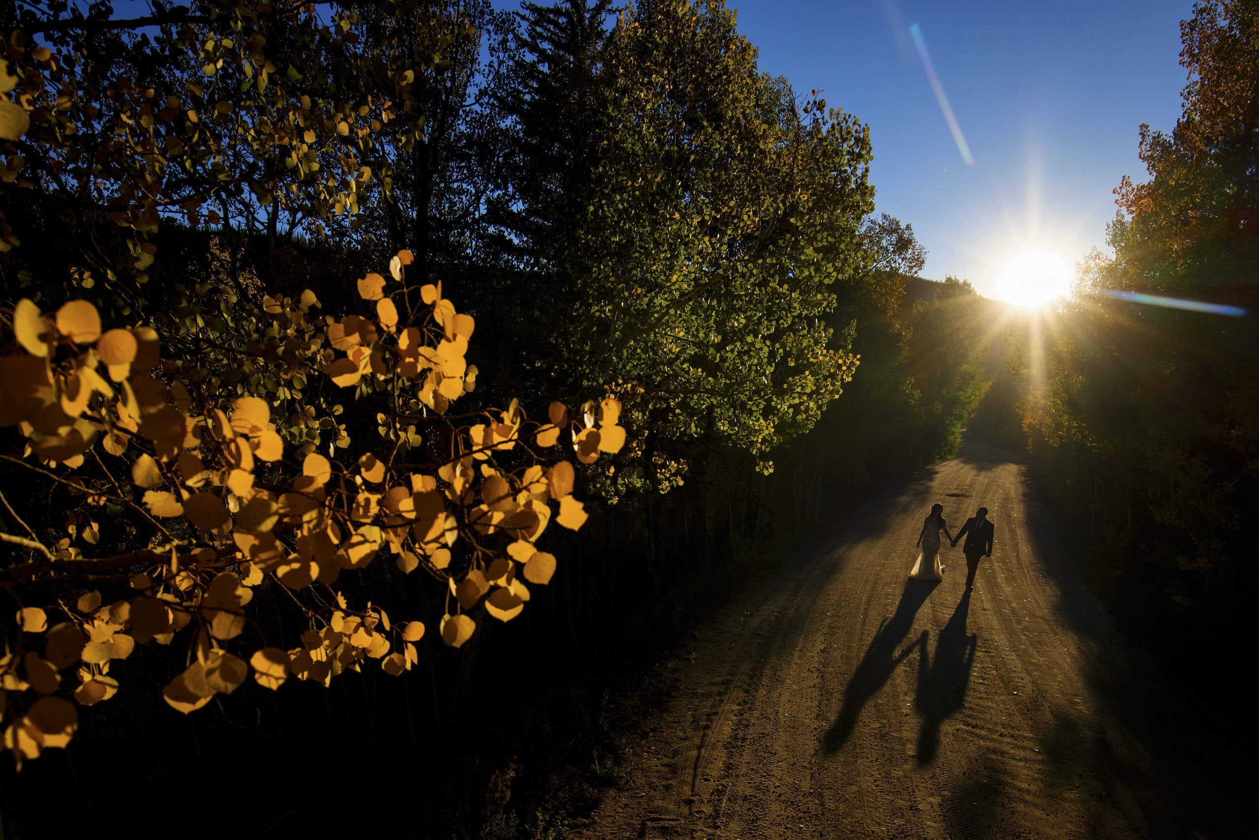 The sun casts an elongated shaddow of newlyweds walking on a dirt road during a fall elopement at Snow Mountain Ranch