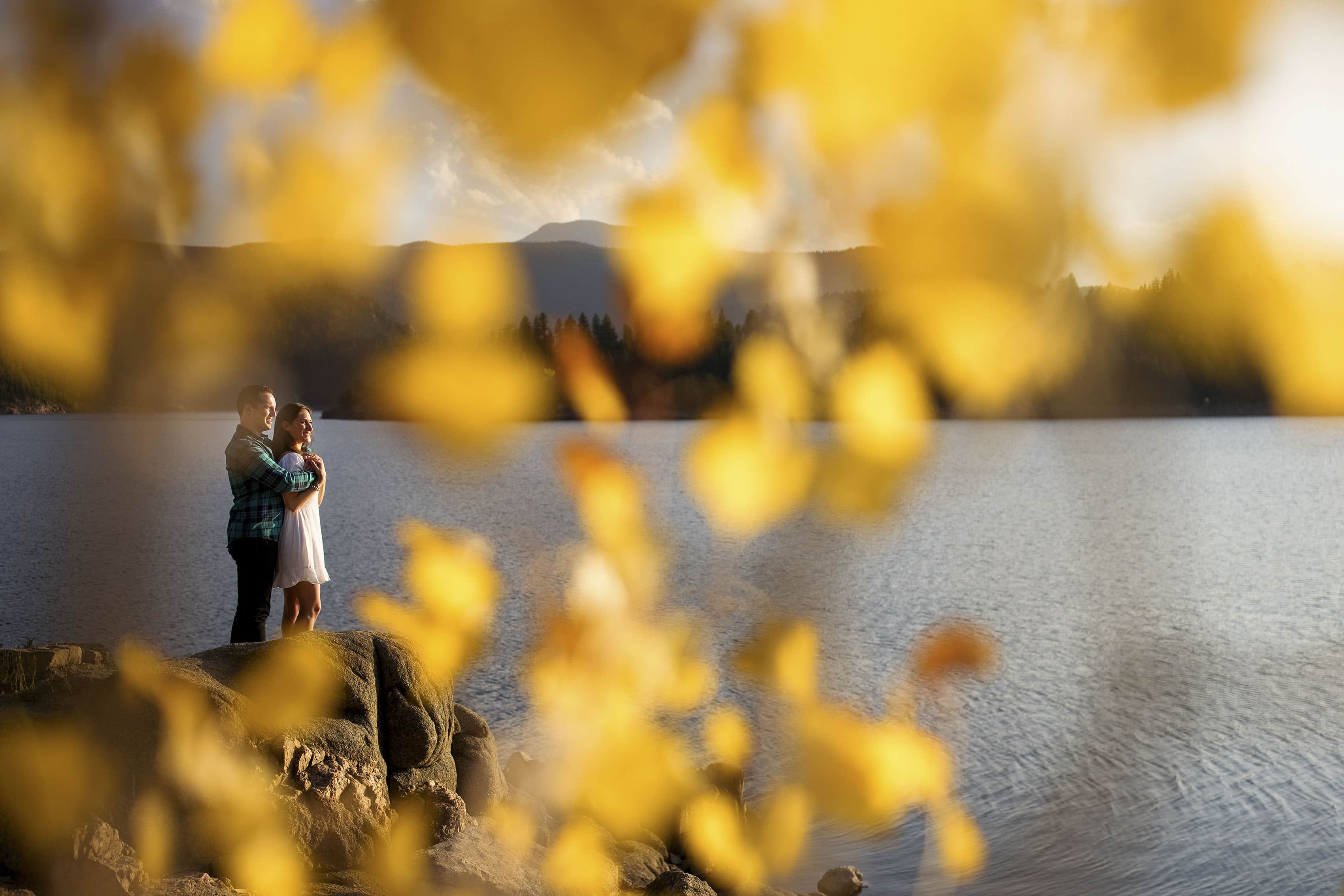 A couple poses near the water’s edge at Gross Reservoir as seen through yellow aspen leaves during their engagement photos