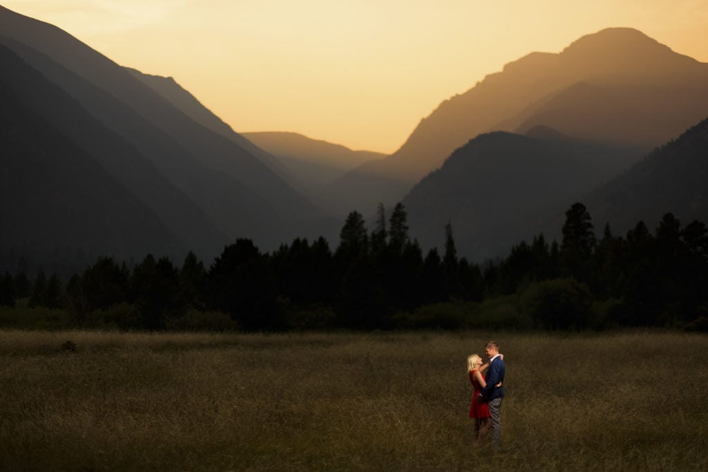 A couple wearing a red dress and blue suit pose in a meadow of tall grass as the sun sets behind the mountains during their engagment pictures in Rocky Mountain National Park