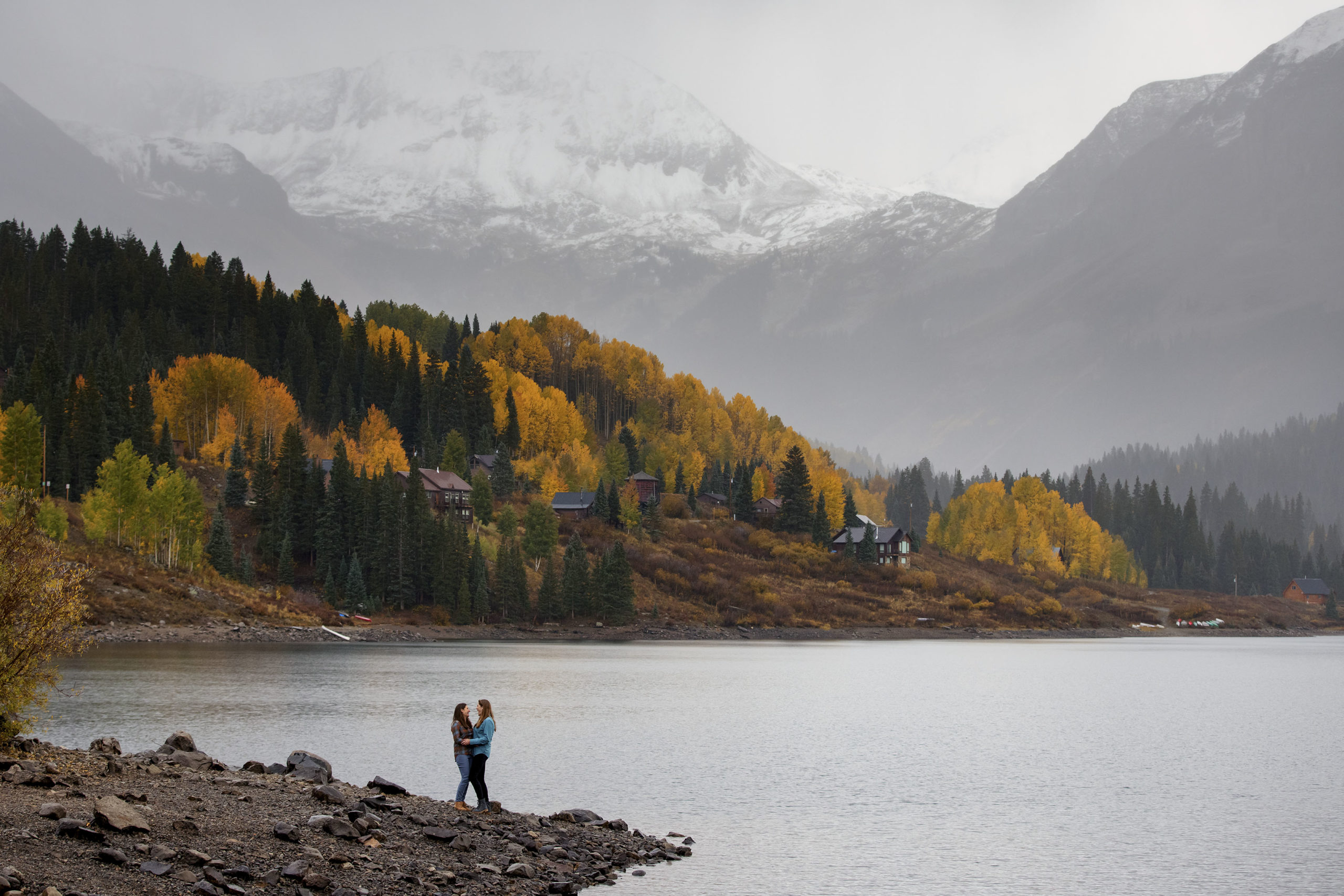 A couple pose on a peninsula at Trout Lake as snow covers the mountain peaks above in Ophir, Colorado during their engagement photos