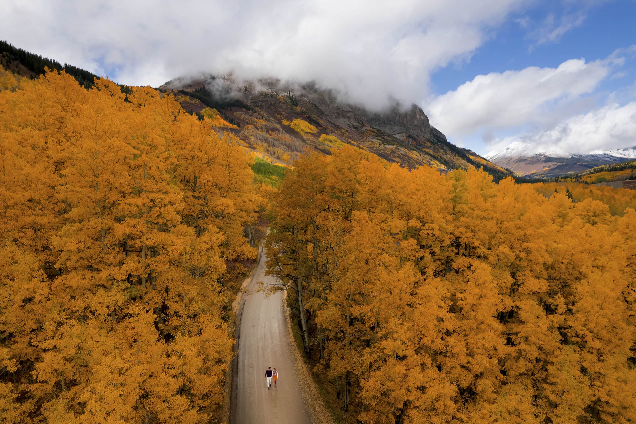 An aerial view of a couple surrounded by golden aspen trees walking along a dirt road in Crested Butte during their engagement session