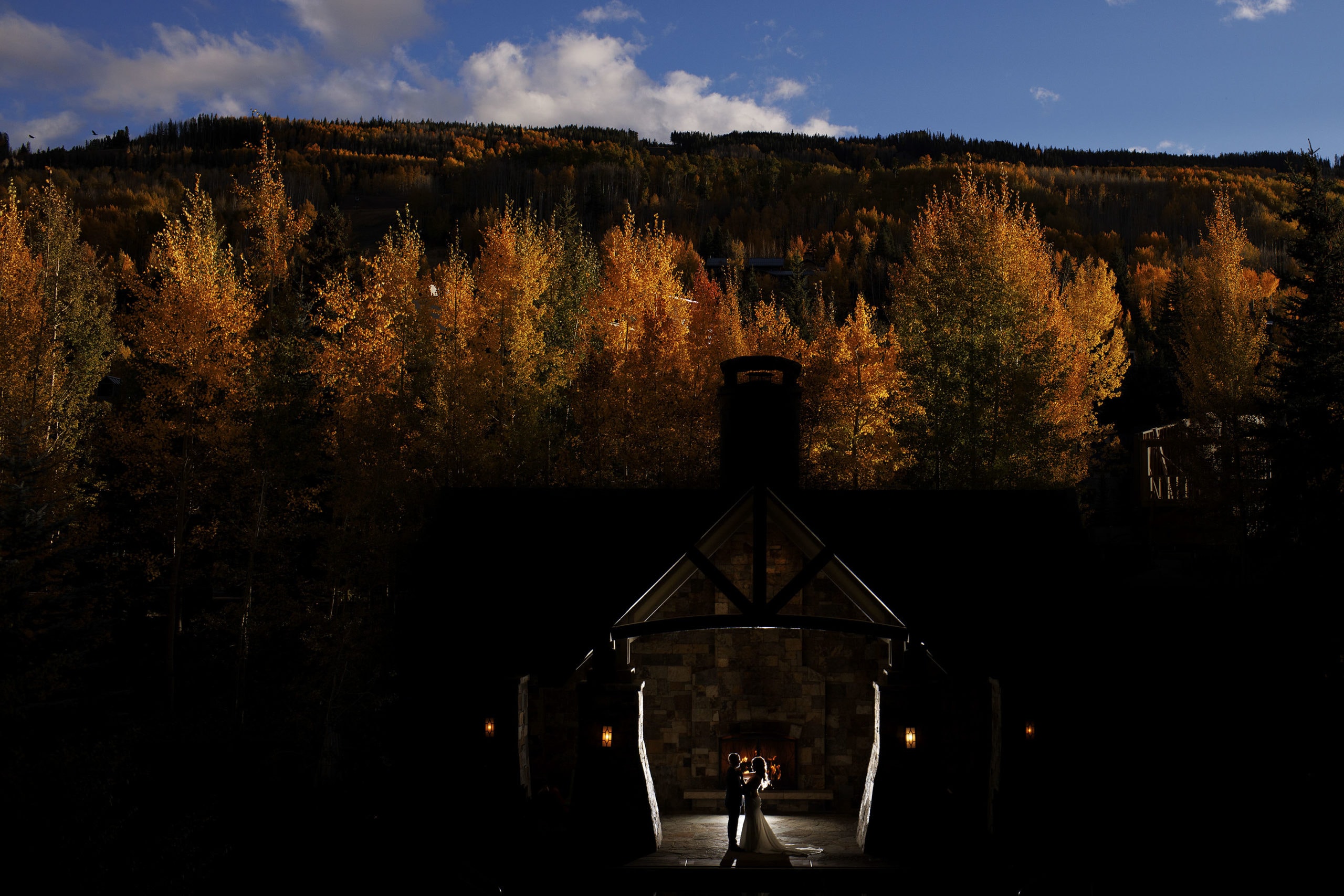 A bride and groom pose in front of the outdoor fireplace at Four Seasons Resort and Residences Vail as the sun illumnates the colorful aspen trees on the hillside