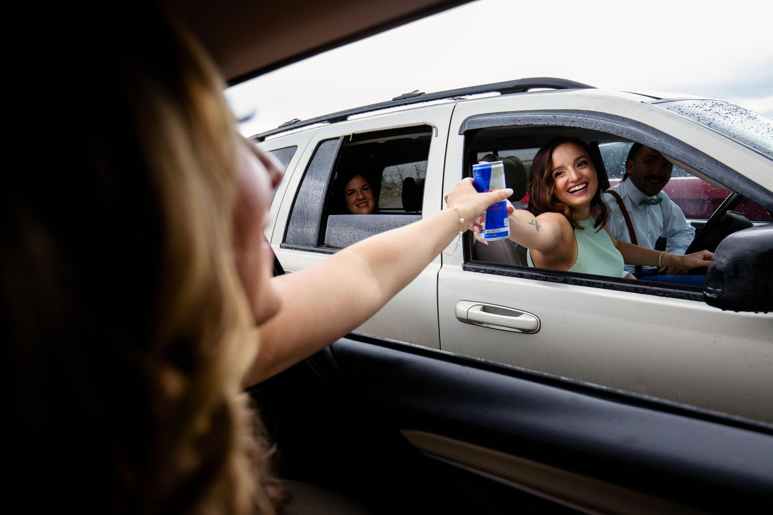 The bride reaches for a can of RedBull from a bridesmaid in the parking lot