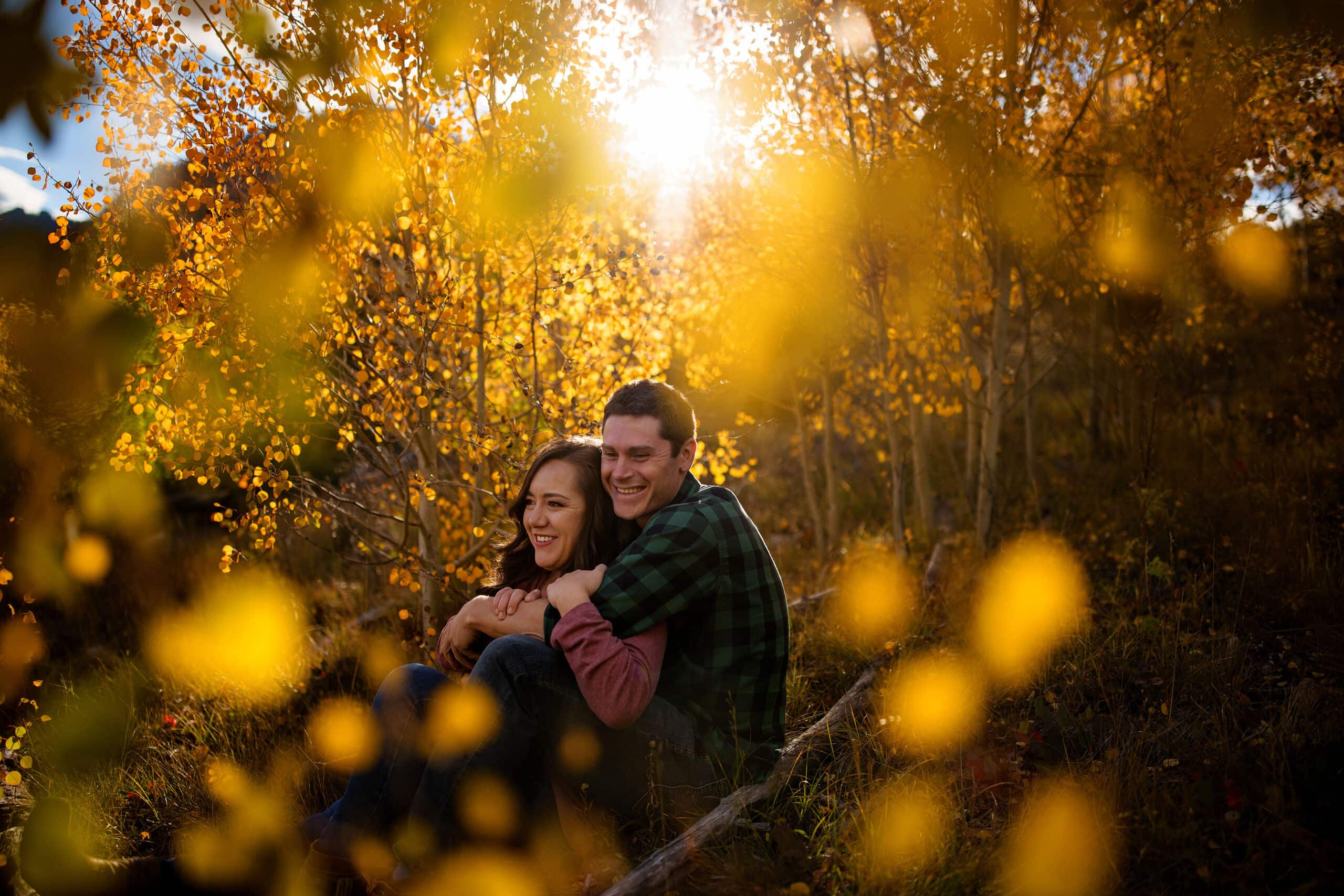 Danielle and Gabe sit on the side of a mountain as the sun shines through the trees behind them