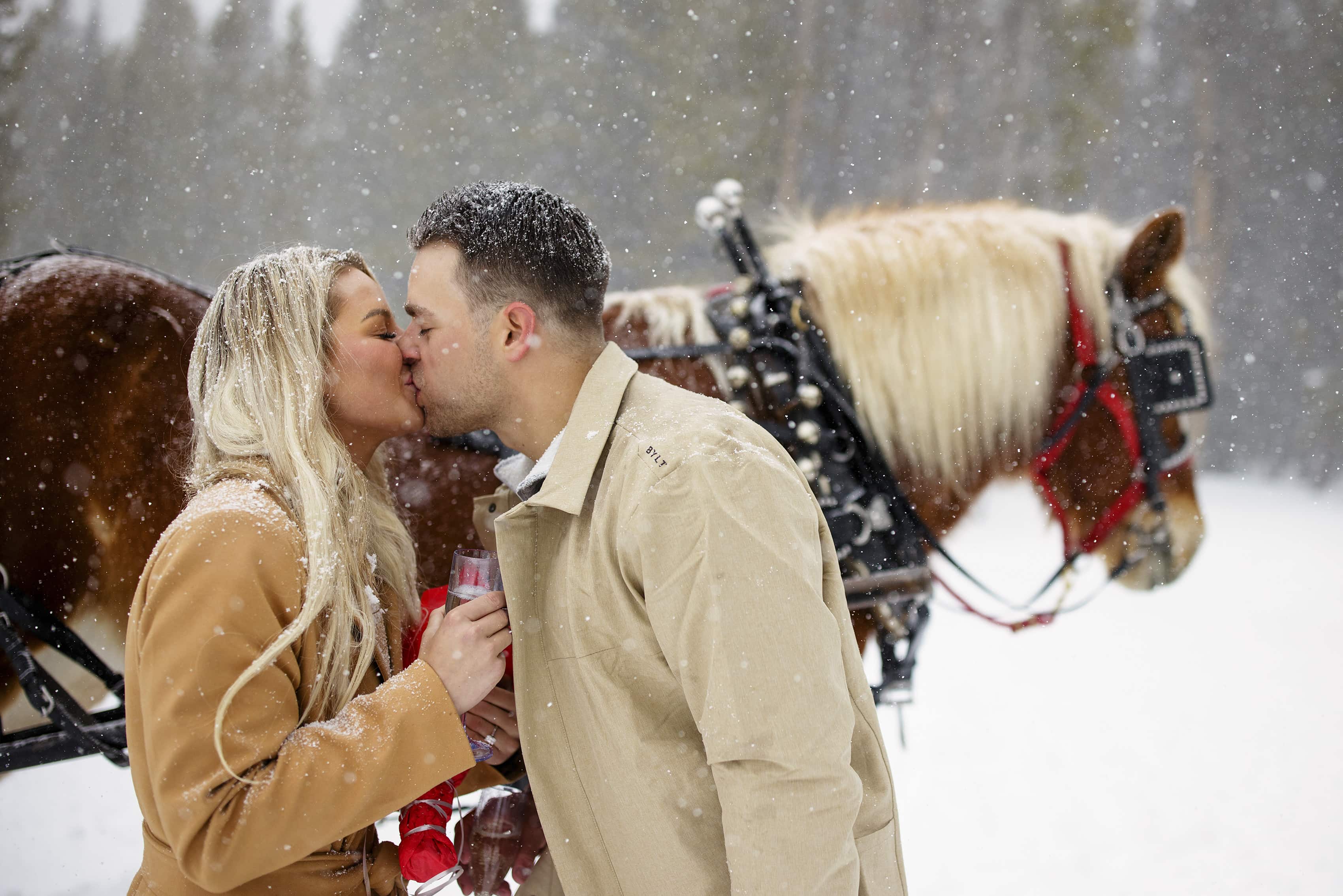 A newly engaged couple kisses near a horse