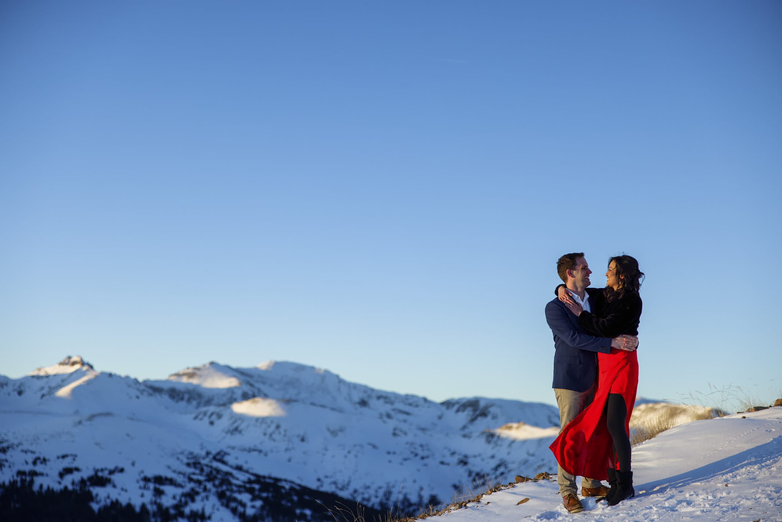 A woman wearing a red dress is held by her spouse in the Rocky Mountains in the snow