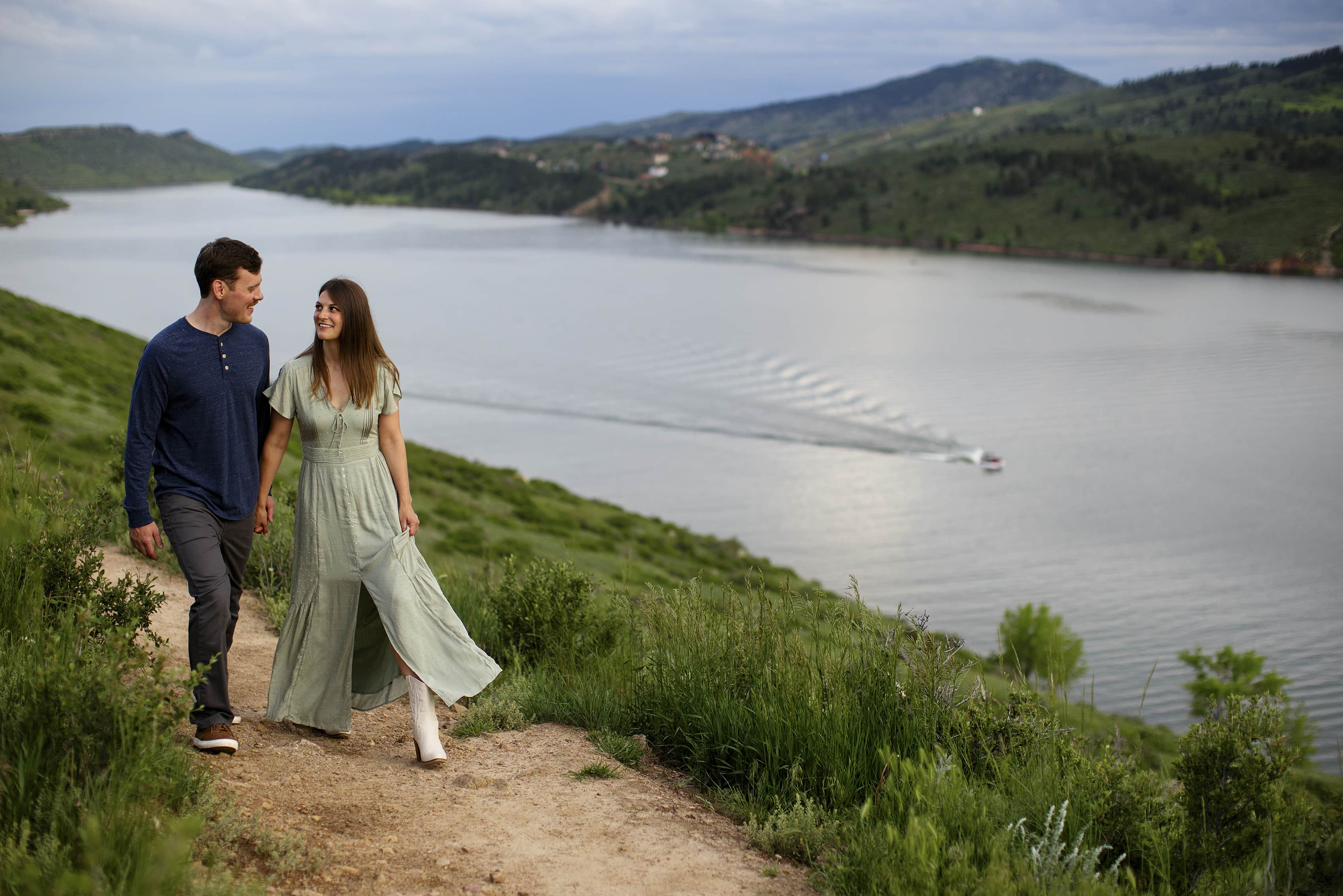 A couple wearing casual clothes walks on a trail as a boat drives by at Horesetooth Reservoir