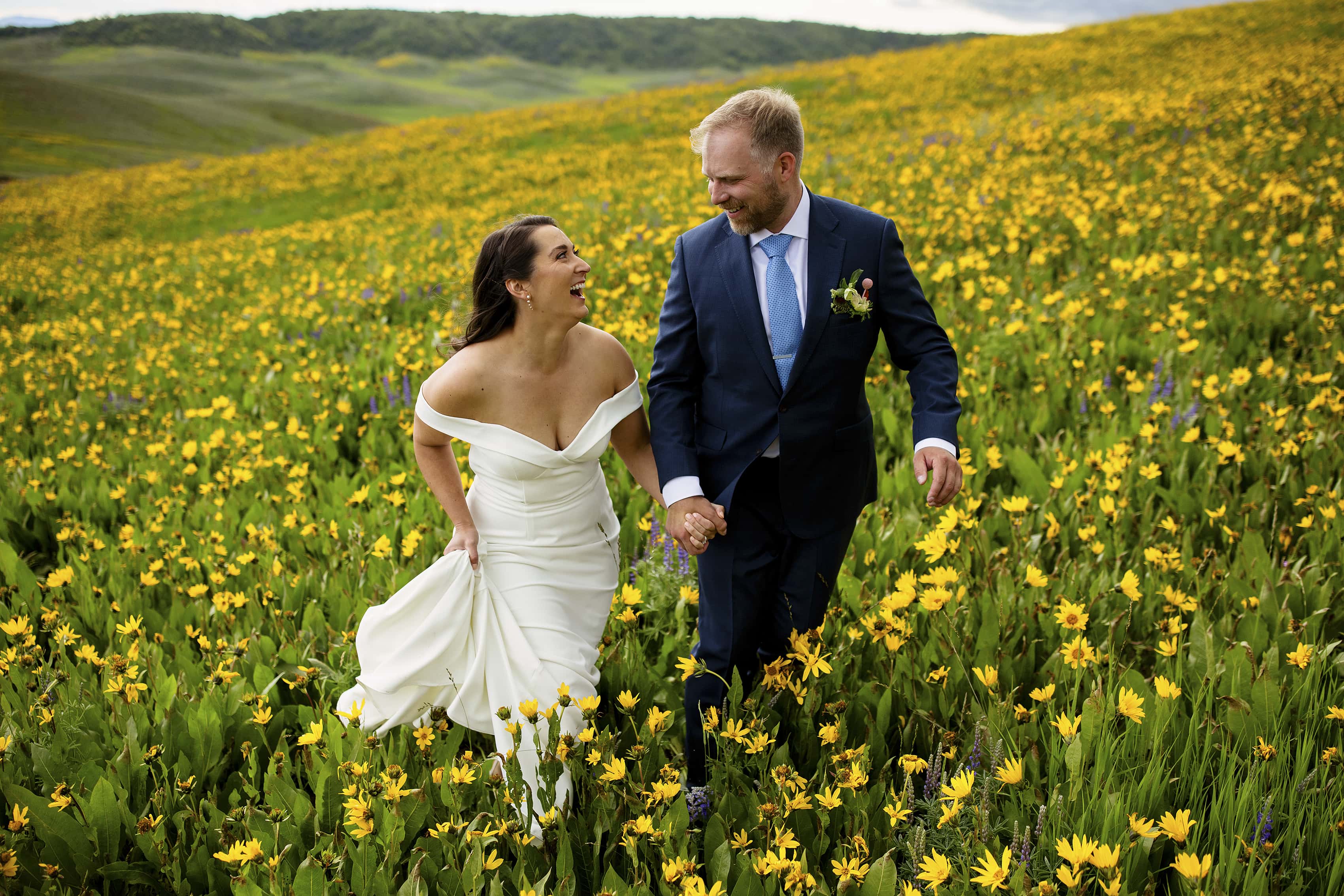 A bride and groom hold hands and walk through a field of daisey and lupine wildflowers in Steamboat Springs