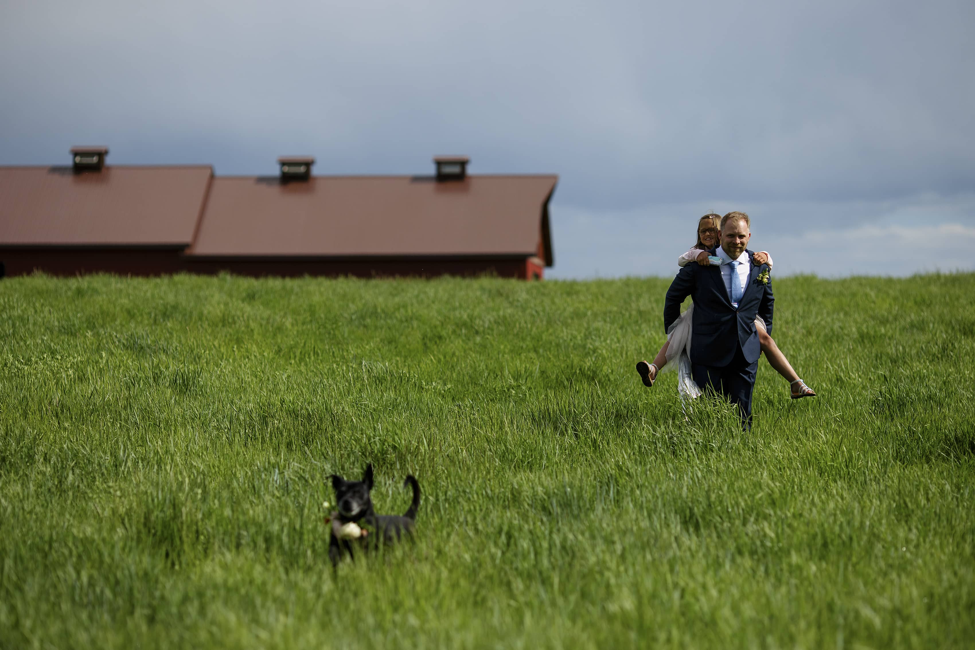 The groom carries his niece through a field as his dog runs ahead