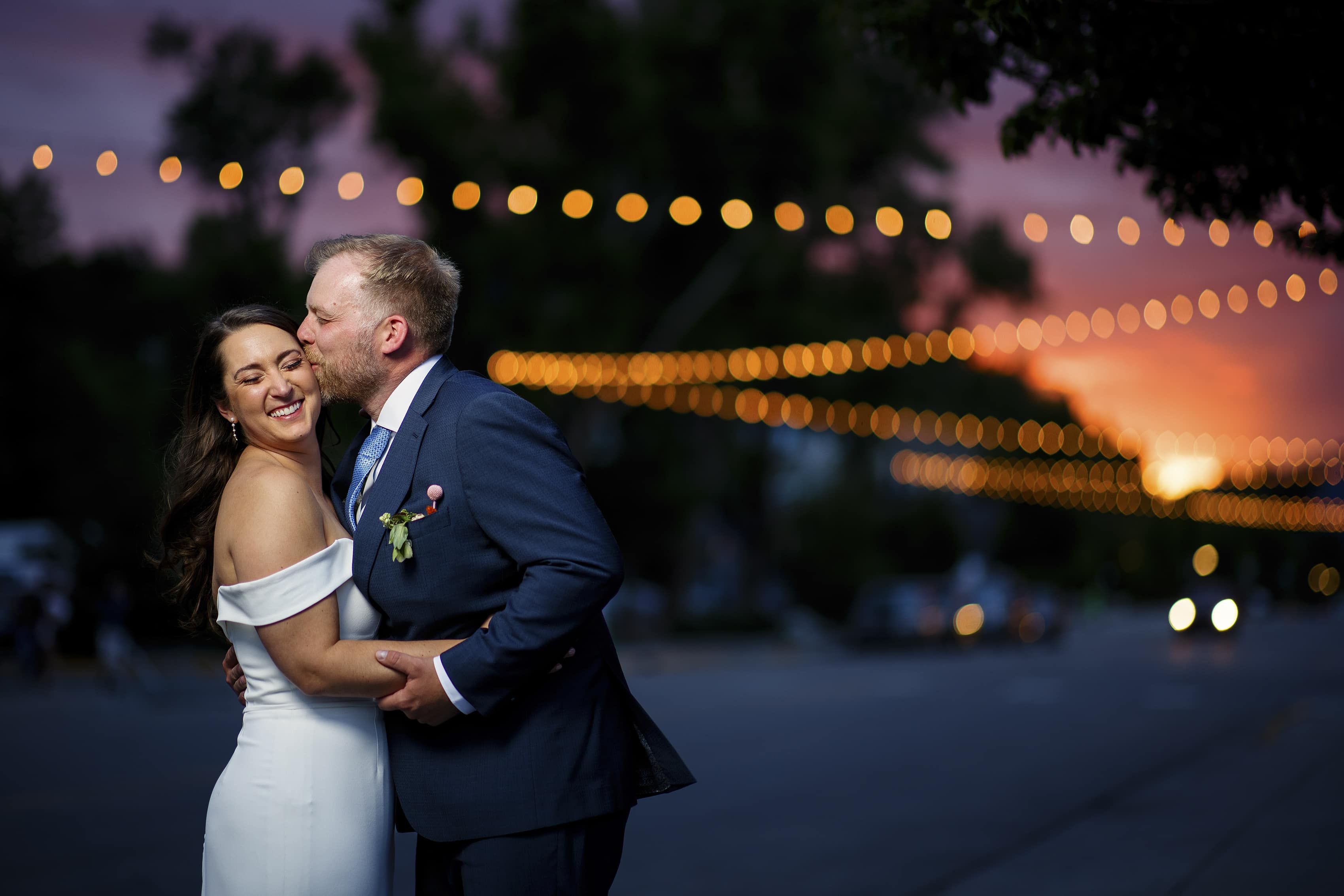 The groom kisses the bride as they pose for a photo on Yampa Street under the market lights at sunset in Steamboat Springs