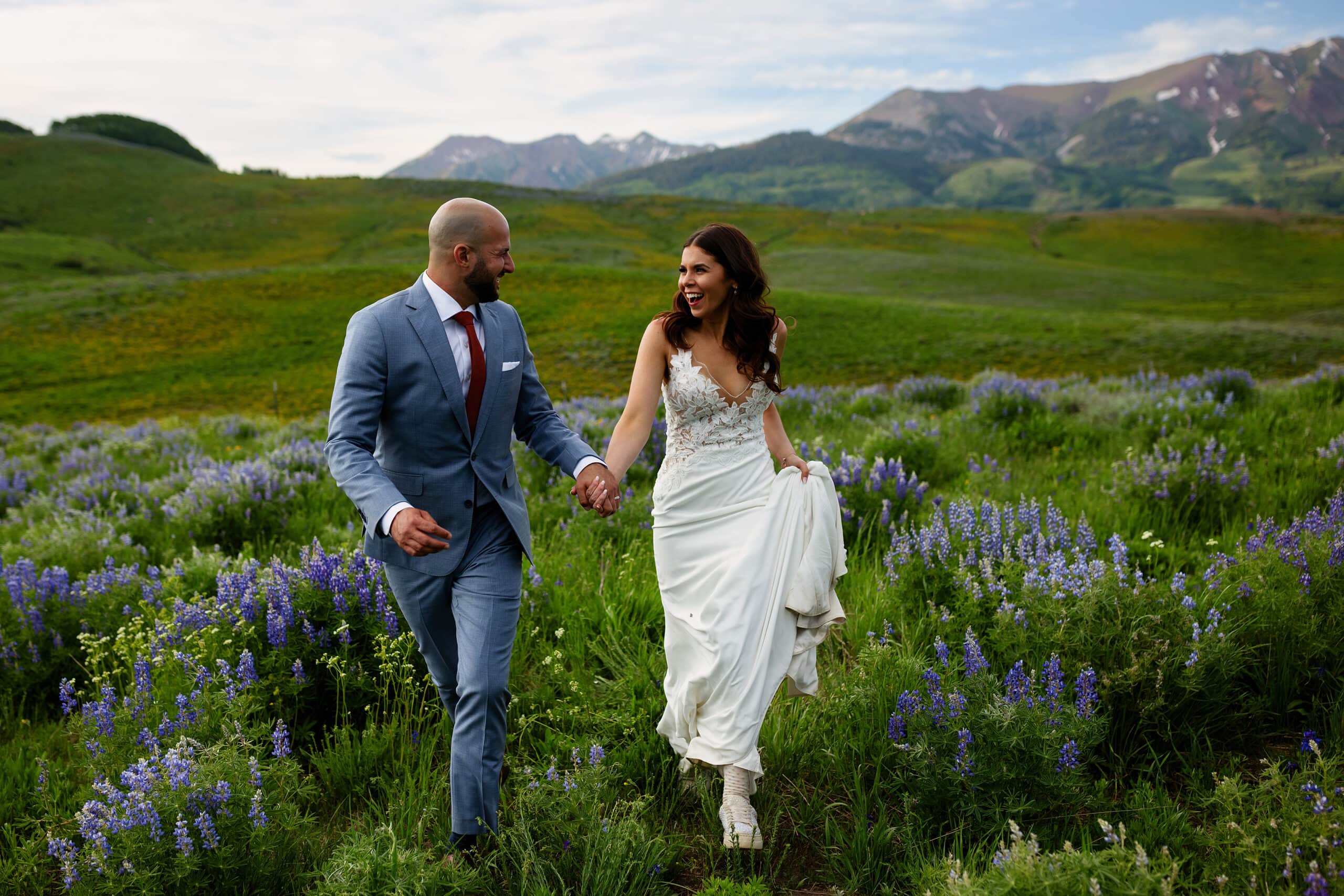 The groom and bride hold hands and run through a field of wildflowers in Crested Butte