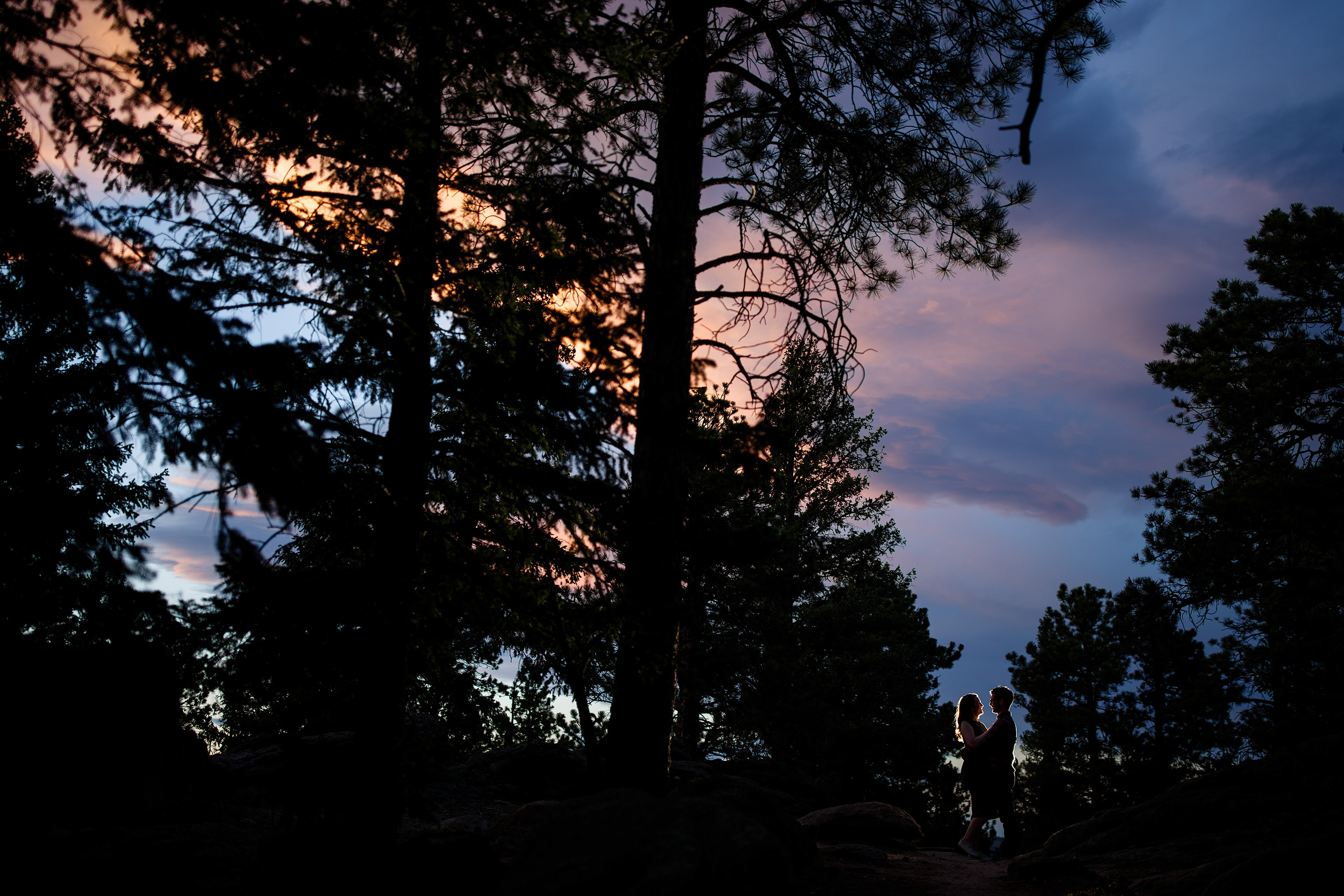 A couple hugs each other as the sun sets behind them in the trees in Evergreen